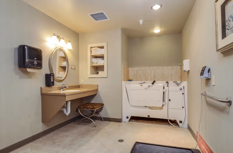 A spacious bathroom with a beige color scheme featuring a wall-mounted sink with a round mirror above it, a cushioned stool underneath, a walk-in bathtub with safety rails, and shelves stocked with folded towels. The room is well-lit with ceiling and wall lights, and has safety features such as grab bars and a soap dispenser.