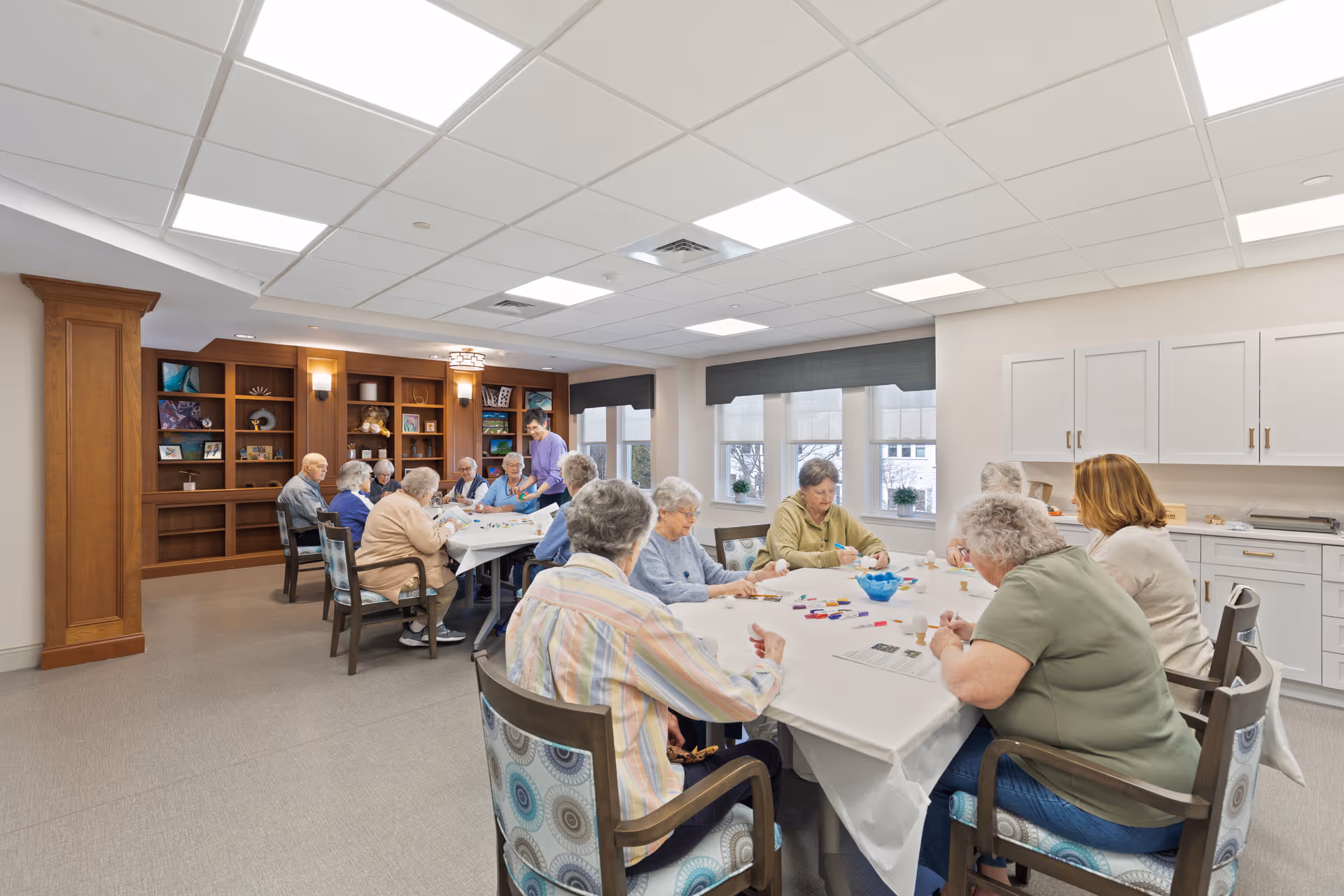 A group of older adults seated around tables in a bright communal activity room doing crafts.