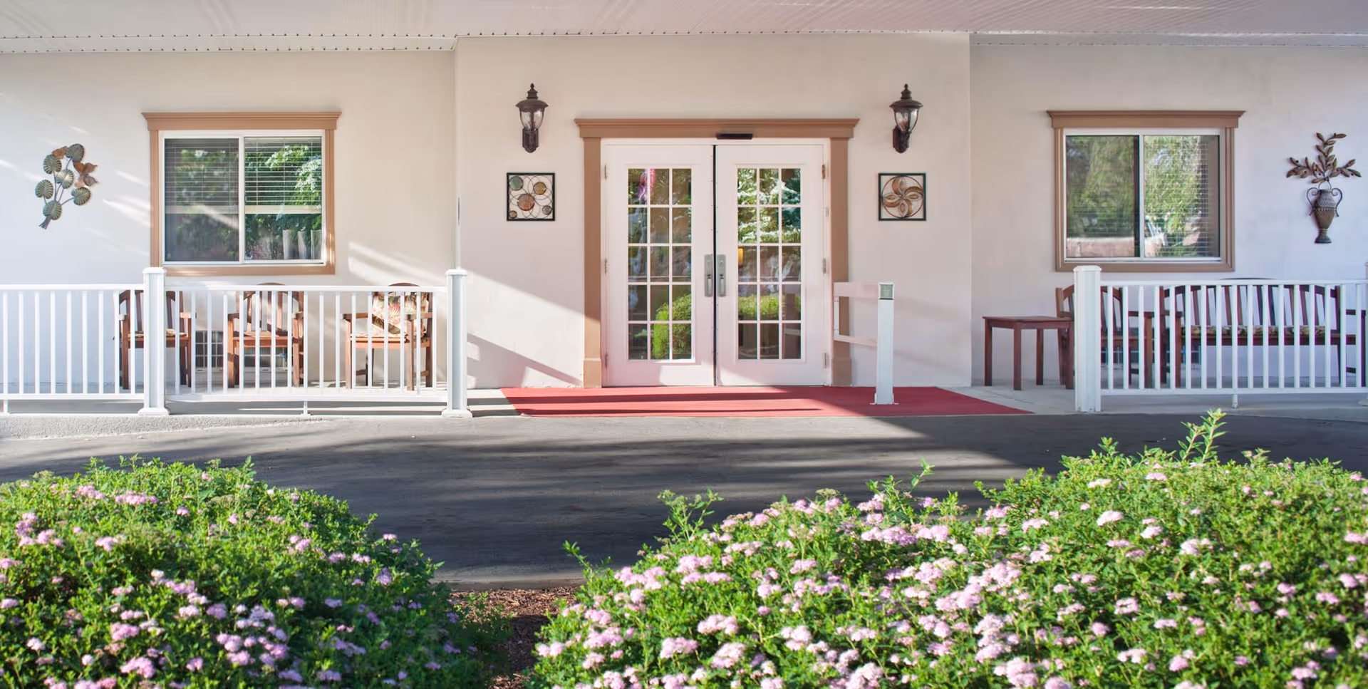 Entrance of a building with double glass doors, flanked by two windows and white railings on either side. There are outdoor benches behind the railings and decorative wall hangings near the doors. In the foreground, there are green bushes with small pink flowers.