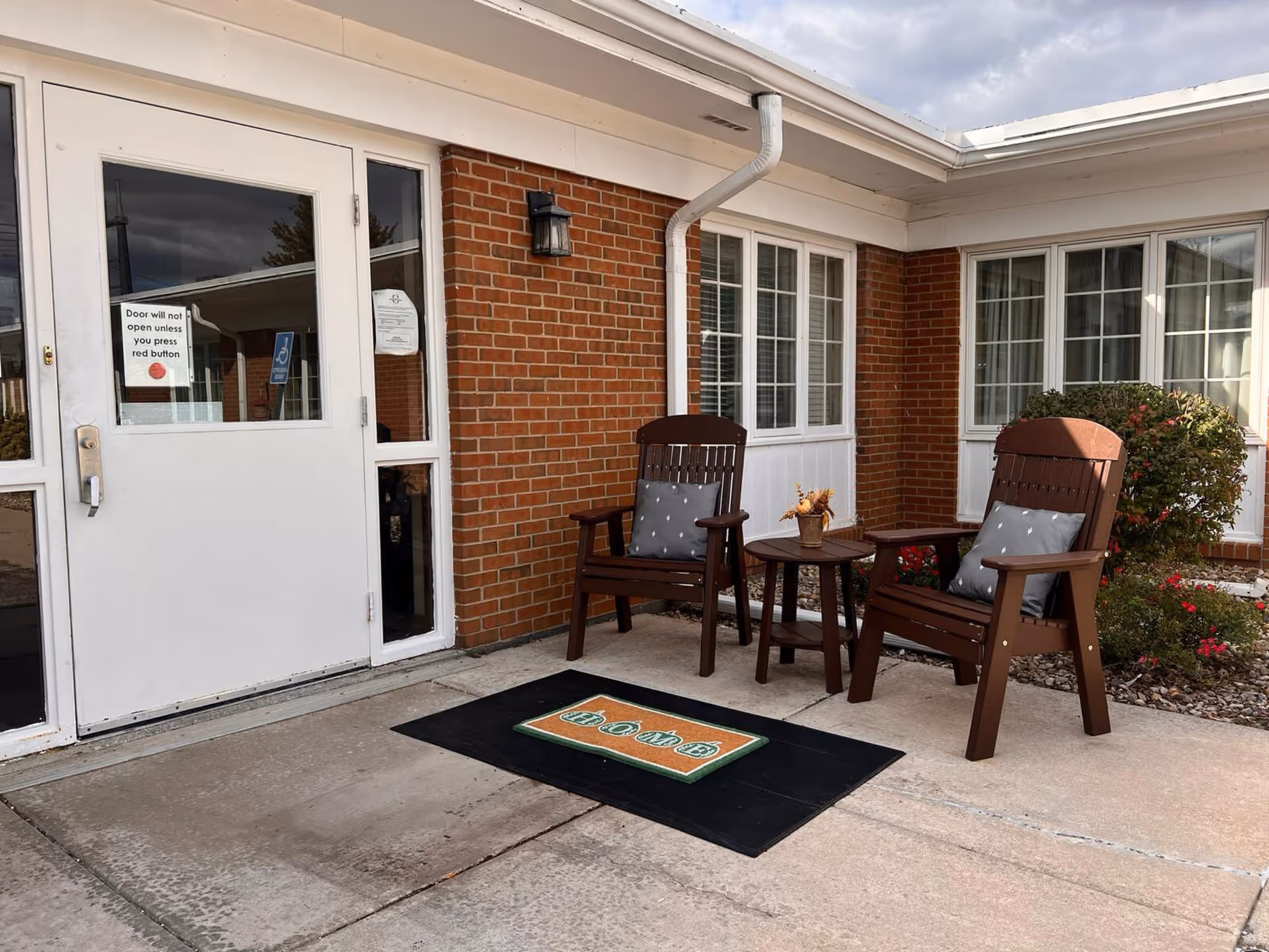 Outdoor seating area at the entrance of a brick building with two wooden chairs with cushions and a small round table between them. A welcome mat is placed on the ground in front of the door. The door has a sign that reads 'Door will not open unless you press red button.'
