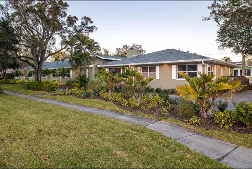 Exterior view of a single-story assisted living facility building surrounded by well-maintained landscaping including palm trees, shrubs, and a green lawn with a sidewalk in front.