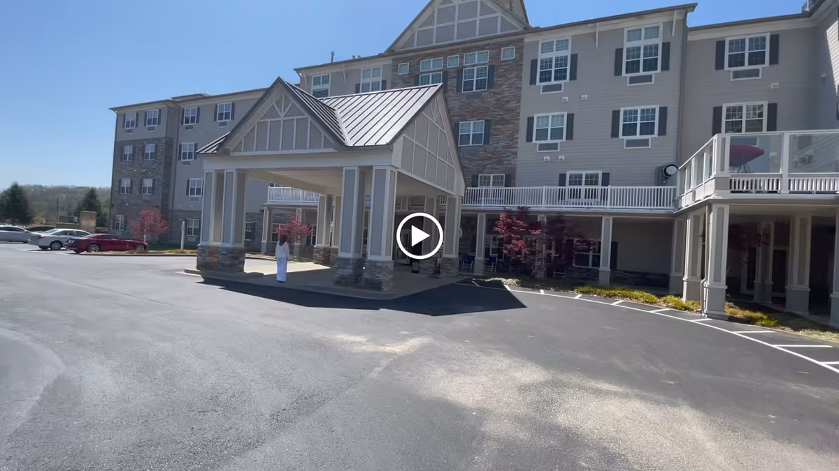 Exterior view of a multi-story senior living facility building with a covered entrance area. Several cars are parked in the parking lot, and a person dressed in white is standing near the entrance. The building features a combination of stone and siding on the facade, with multiple windows and balconies. The sky is clear and blue.