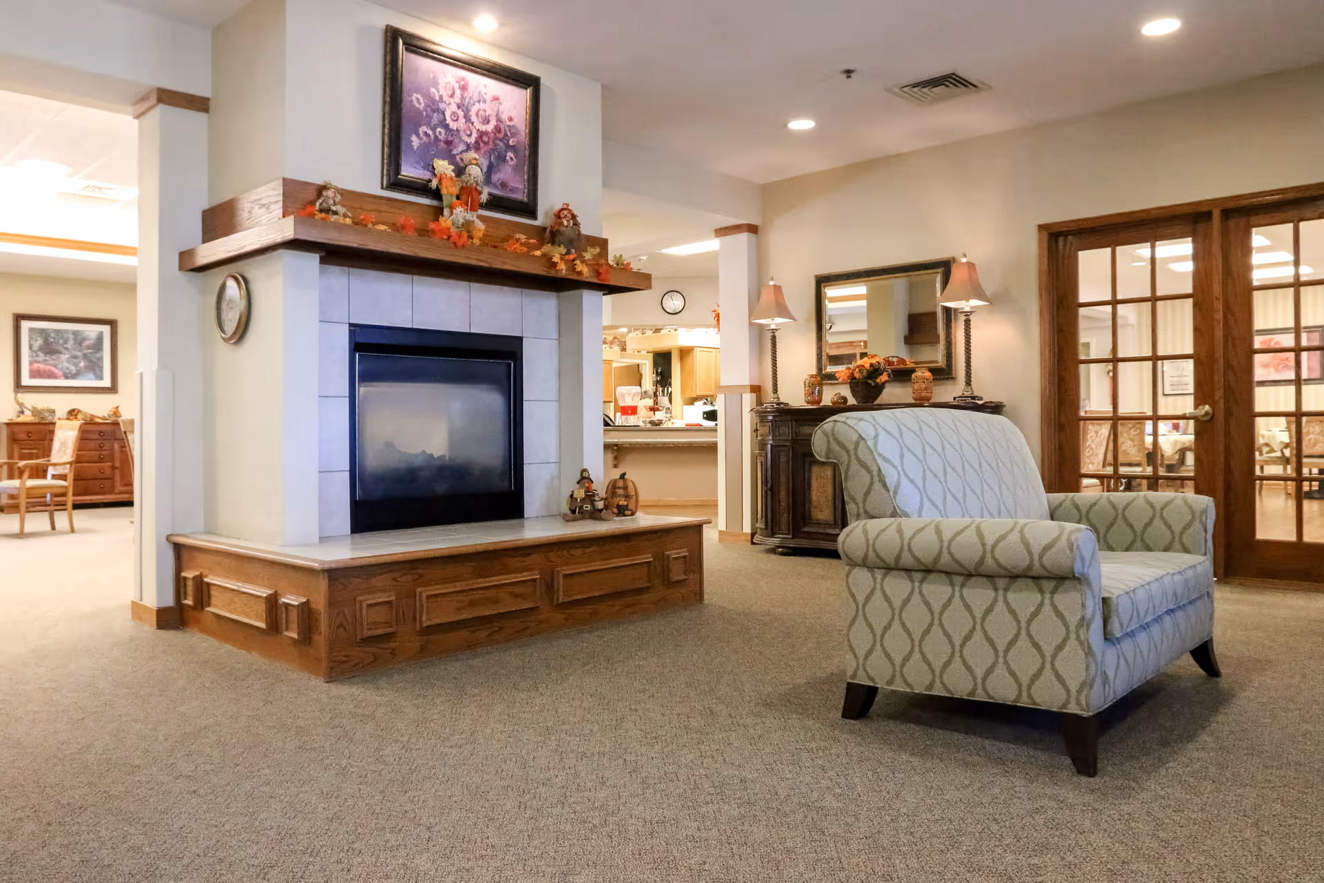 A cozy interior common area featuring a fireplace with a wooden mantle decorated with autumn-themed ornaments and a framed floral painting above it. In front of the fireplace is a patterned armchair. To the right, there is a wooden sideboard with two lamps and a mirror above it. The room has beige walls, carpeted floors, and wooden framed glass double doors leading to another room with chairs and tables.