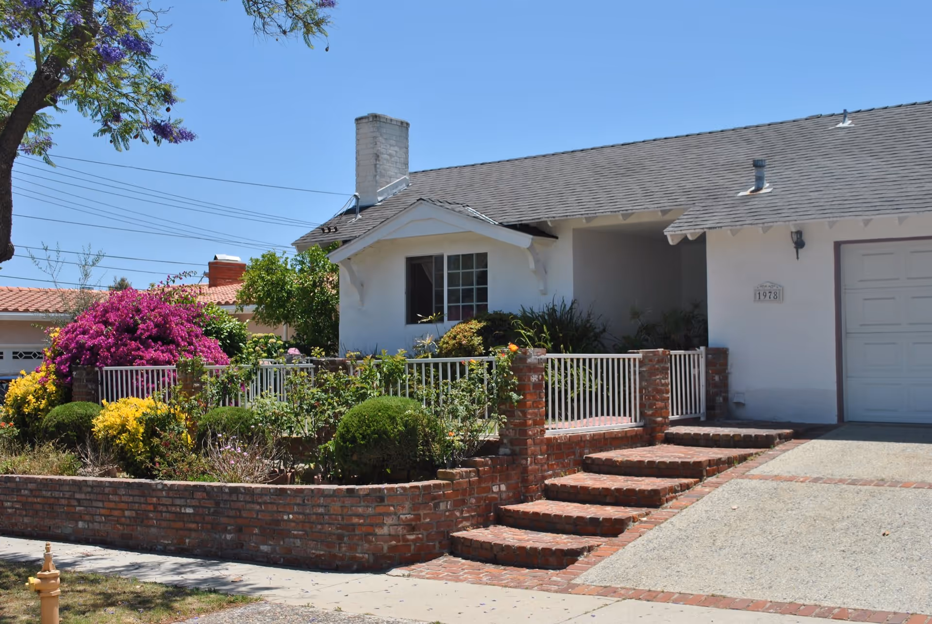 Front exterior view of a single-story white house with a gray shingled roof, a brick staircase leading to the entrance, a small garden with various green plants and colorful flowers, and a driveway next to the garage door.