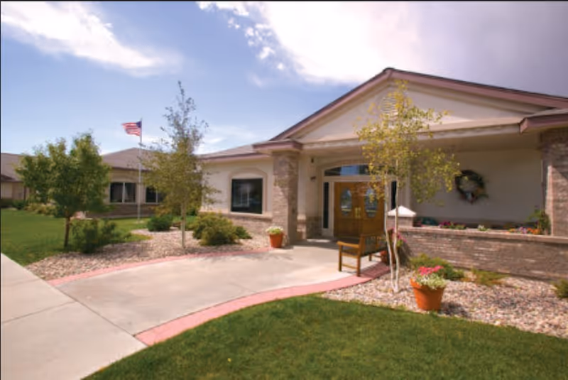 Exterior view of a single-story senior living facility building with a covered entrance, wooden double doors, a bench, potted plants, small trees, and an American flag on a flagpole. The sky is partly cloudy.