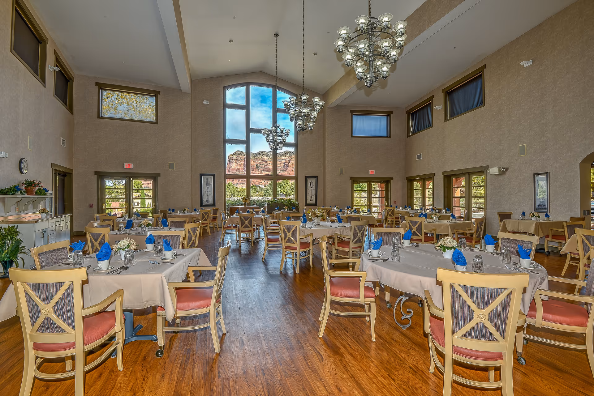 A spacious dining room with multiple tables covered in beige tablecloths, each set with blue napkins, cups, glasses, and silverware. The room features large windows with a scenic view of red rock formations and greenery outside. The walls are beige, and there are several chandeliers hanging from the high ceiling. Wooden chairs with red cushions surround the tables, and there are plants and framed artwork on the walls.