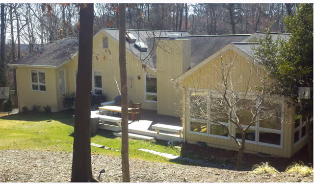 Exterior view of a yellow single-story building with multiple windows and a small wooden deck with benches, surrounded by trees and grass.