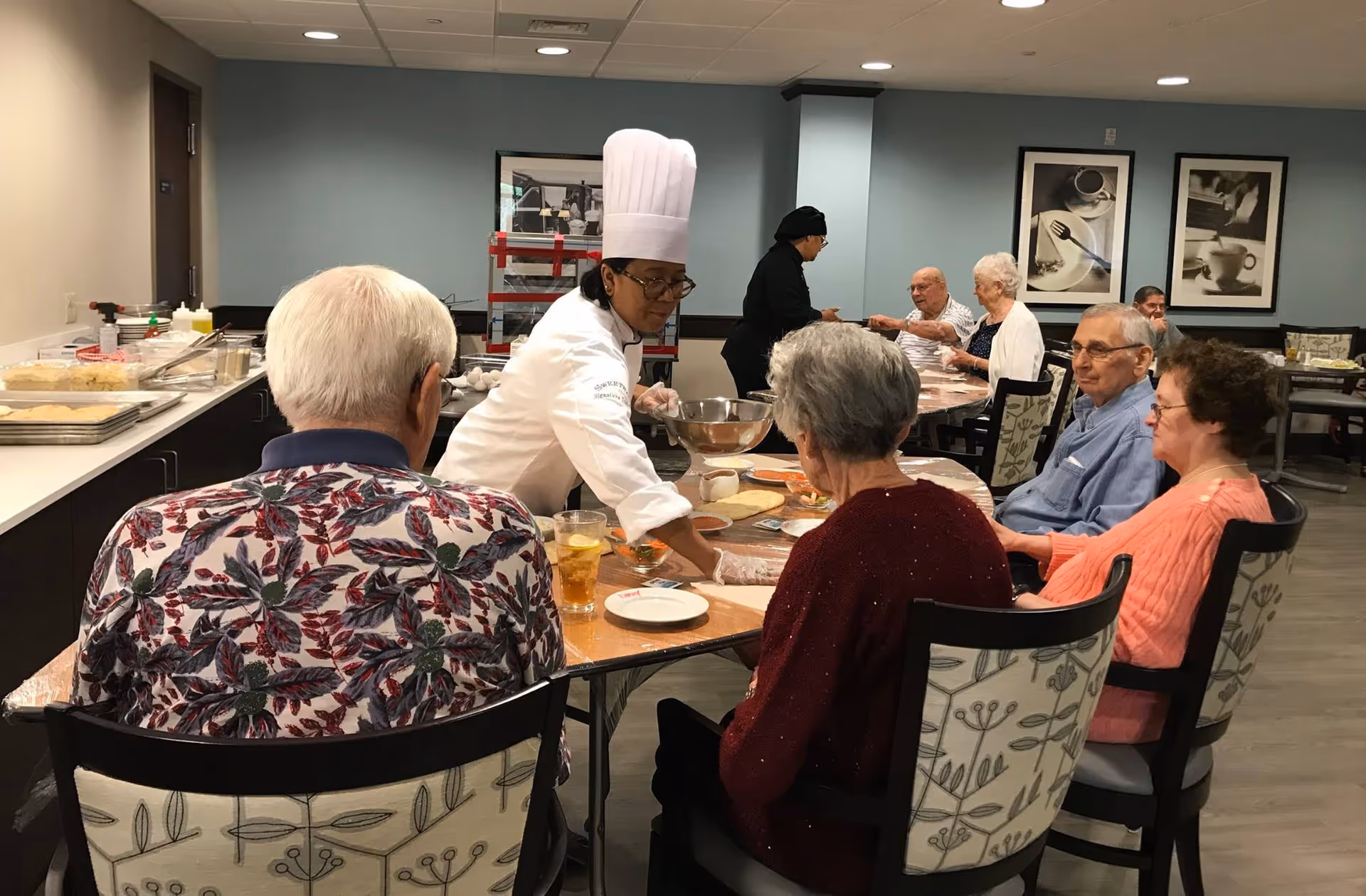 A chef wearing a white uniform and tall chef hat is serving food to a group of elderly people seated around a dining table in a communal dining area. The room has light blue walls with framed pictures of coffee cups and utensils. Several elderly individuals are seated and engaged in the meal or conversation.