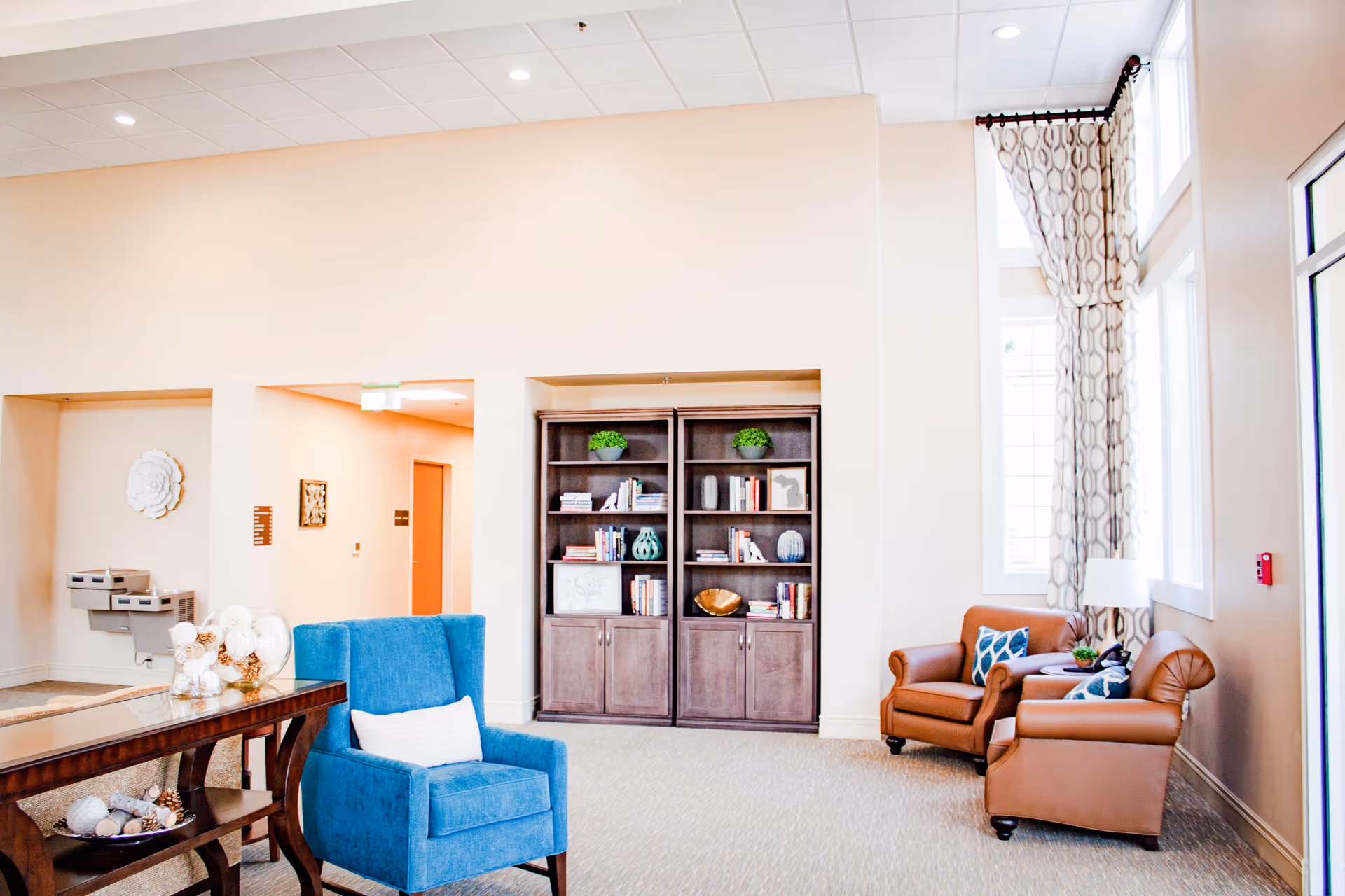 A bright and spacious living room area in a senior living facility with a blue armchair, two brown leather armchairs with decorative pillows, a wooden bookshelf filled with books and decorative items, large windows with patterned curtains, and a wooden table with decorative objects.