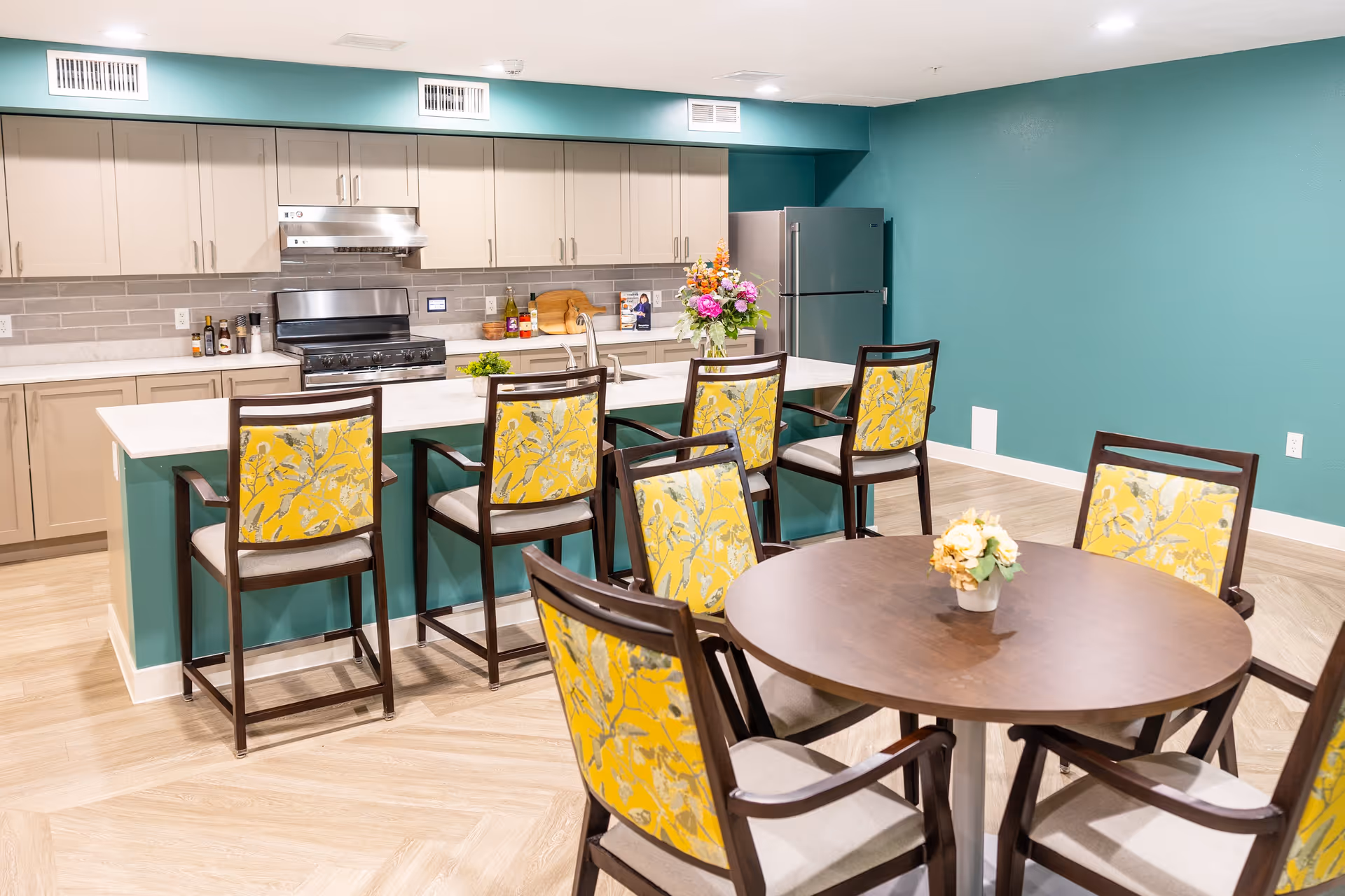 A modern kitchen and dining area in a senior living facility. The kitchen features beige cabinets, a stainless steel stove and refrigerator, and a white countertop island with a sink. Five high chairs with yellow floral upholstery are placed at the island. In the foreground, there is a round wooden dining table surrounded by six chairs with matching yellow floral upholstery. A small flower arrangement is on the dining table. The walls are painted teal, and the floor has a light wood pattern.
