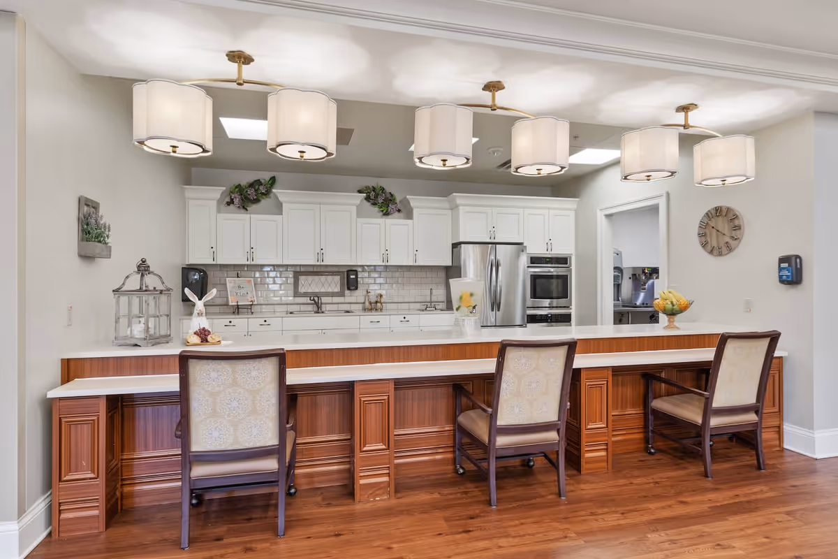 A bright and clean kitchen area with white cabinets, stainless steel refrigerator and oven, and a long wooden counter with three upholstered chairs. The counter has decorative items including a glass container with water and lemon slices, a bowl of bananas, and a small bunny figurine. Above the counter are five modern pendant lights with white shades. The floor is wooden, and there is a wall clock and a hand sanitizer dispenser on the right wall.