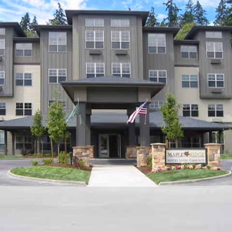 Front exterior view of a multi-story senior living facility named Maple Ridge by Bonaventure, featuring a covered entrance with two flags and landscaped greenery.