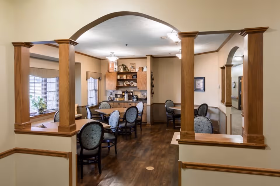 Interior view of a dining area in a senior living facility with wooden columns and archways. The room features multiple round tables with cushioned chairs, wood flooring, and windows with blinds. A small kitchenette area with cabinets, a coffee maker, and decorative items is visible in the background.