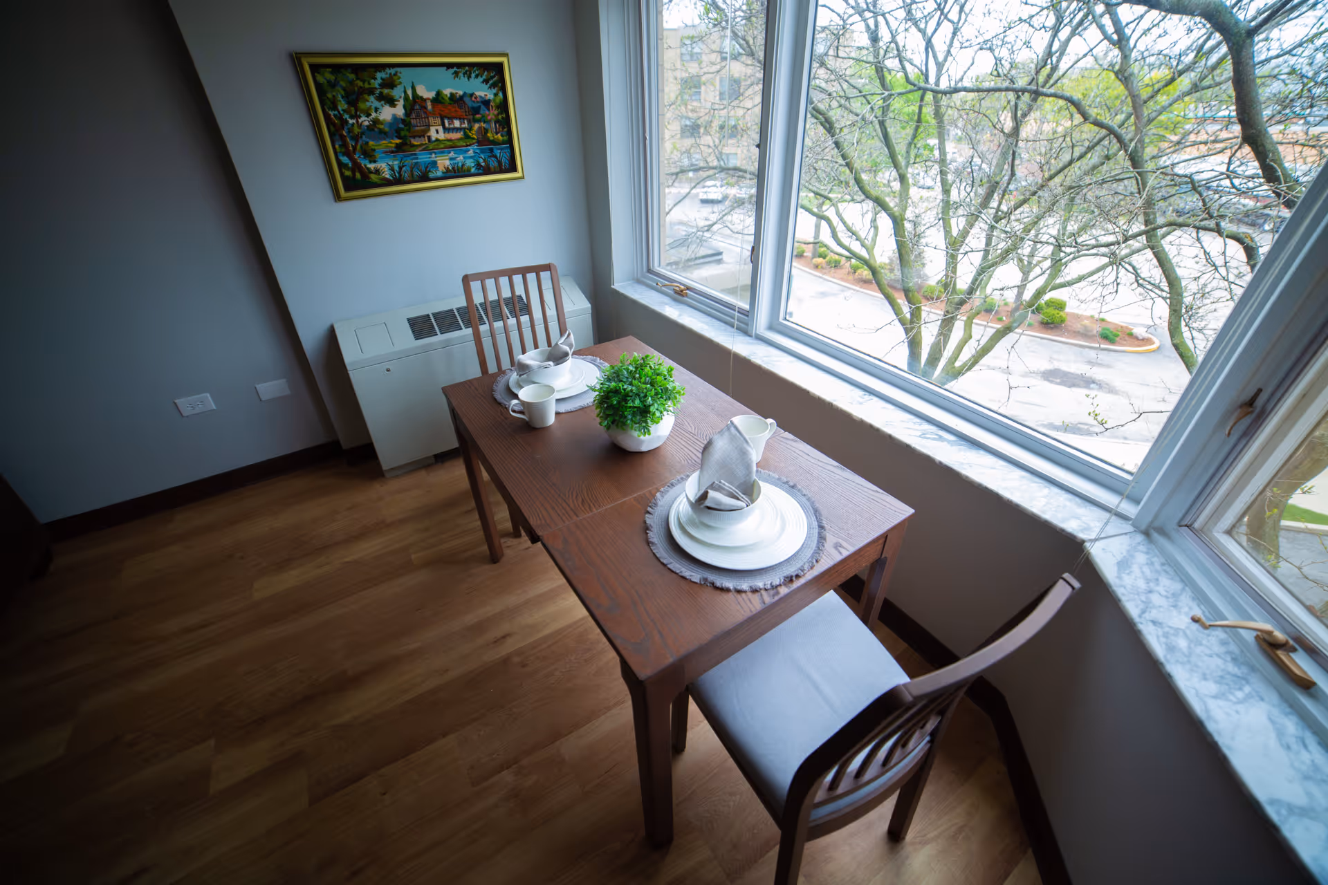 A small dining area with a wooden table set for two, including plates, cups, and napkins. There is a small green plant centerpiece on the table. Two wooden chairs with cushions are placed on either side of the table. The dining area is next to large windows showing a view of trees and a parking lot outside. A framed painting hangs on the wall above a white radiator cover.