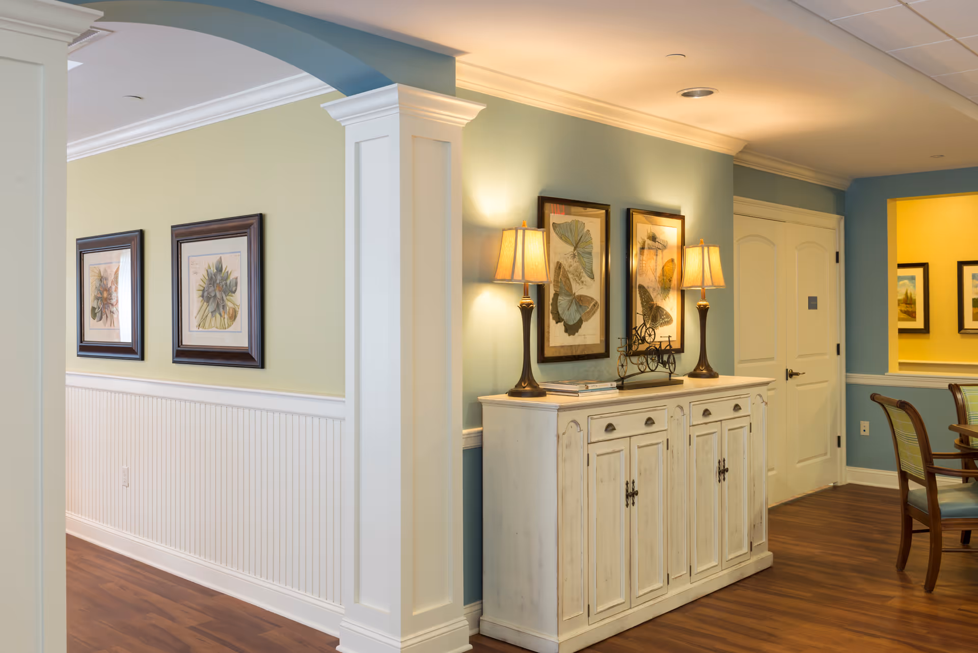 Interior view of a senior living facility hallway and common area with light blue and yellow walls, wooden flooring, a white sideboard with two table lamps, framed butterfly artwork above the sideboard, and a dining area with chairs visible to the right.