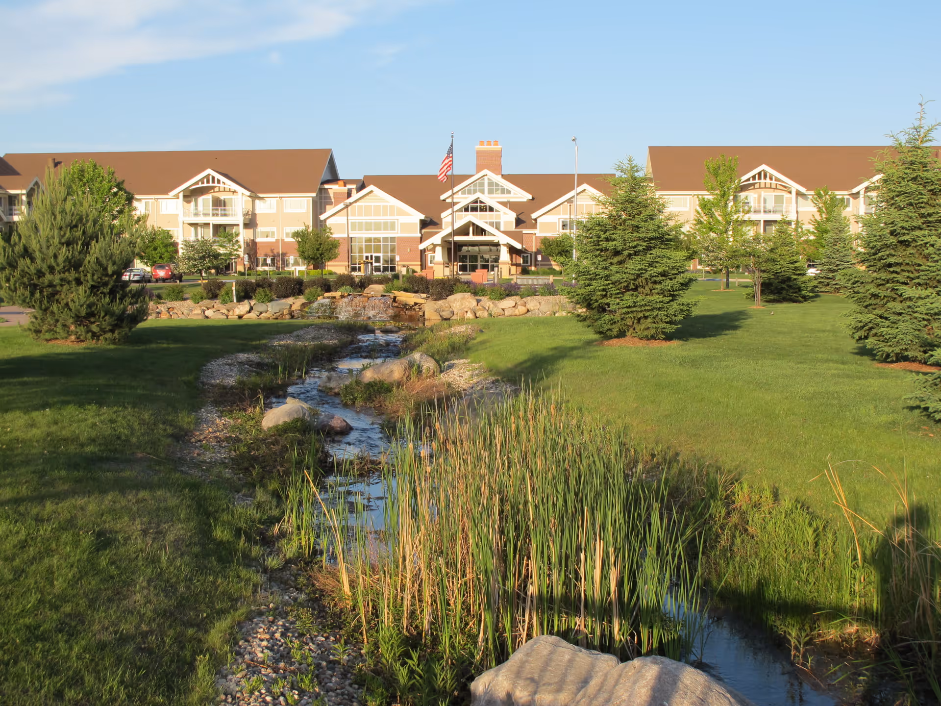 View of the Good Samaritan Society - Prairie Creek facility with a landscaped garden in the foreground featuring a small stream, rocks, green grass, and several trees under a clear blue sky.