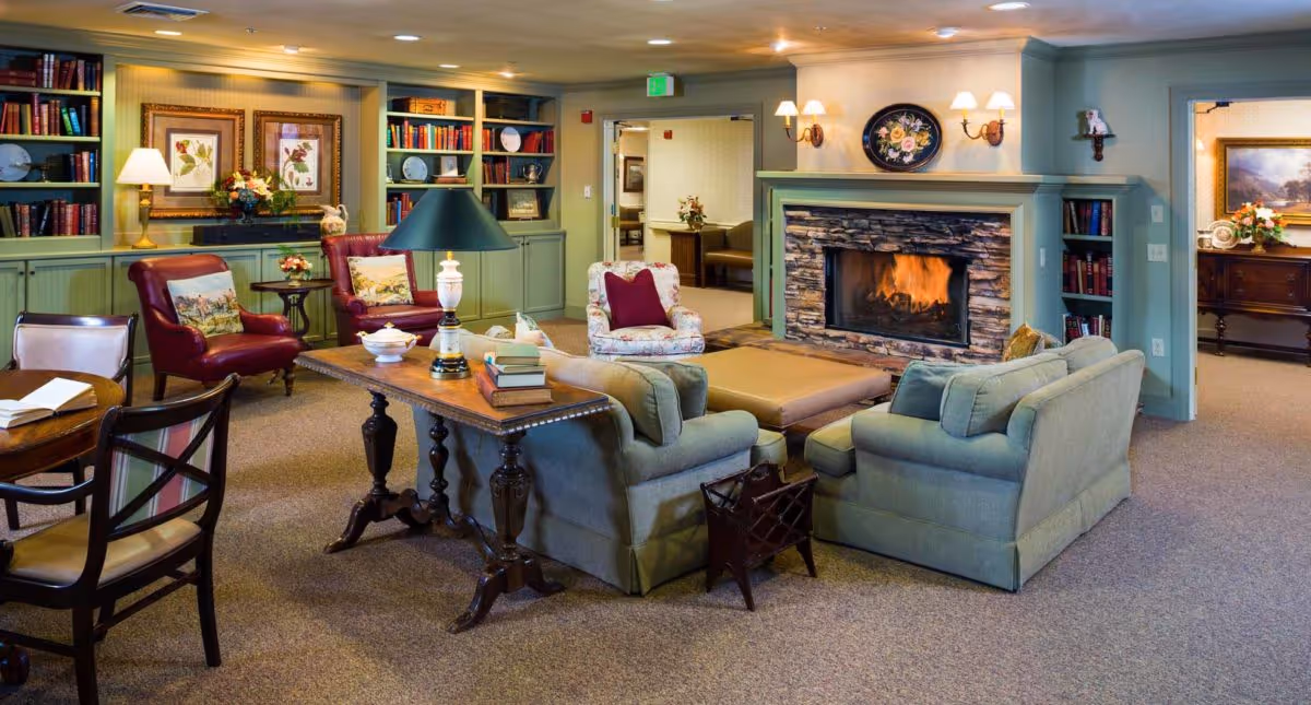 A cozy living room in an assisted living community featuring a stone fireplace with a fire burning, surrounded by comfortable seating including two green sofas, a floral armchair, and two red leather chairs. The room has built-in green bookshelves filled with books and decorative items, a wooden table with a lamp and books, and soft lighting from wall sconces and a table lamp. There is a carpeted floor and artwork on the walls, creating a warm and inviting atmosphere.