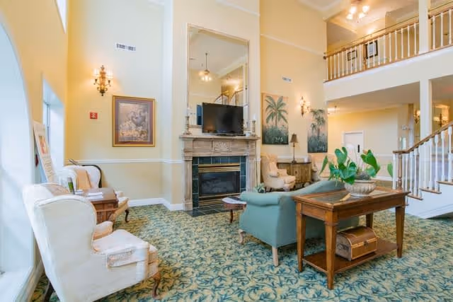 A spacious and well-lit living room area with high ceilings, featuring a fireplace with a mounted TV above it, comfortable armchairs, a green sofa, a wooden table with a plant, and decorative paintings on the walls. There is a staircase with white railings leading to an upper level.