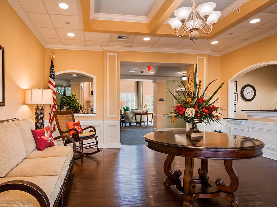 A warmly lit senior living facility common area with beige walls and dark wooden flooring. There is a beige sofa with red decorative pillows and a wooden rocking chair with a red cushion on the left. A round wooden table with a large floral arrangement is centered in the foreground. The background shows an open doorway leading to another seating area with chairs and tables, and an American flag is positioned near the sofa. A chandelier hangs from the ceiling, and a clock is visible on the wall to the right.