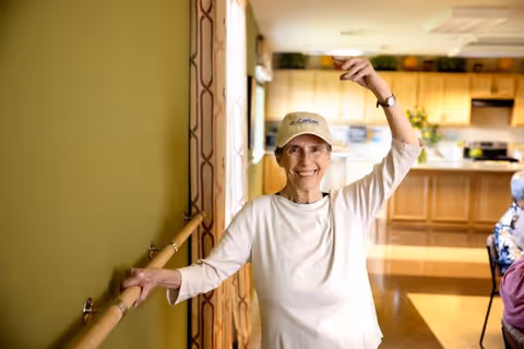 An elderly woman wearing a beige hat and light-colored long sleeve shirt is smiling and holding onto a handrail on a green wall while raising one arm. She is standing in a well-lit room with a kitchen visible in the background.