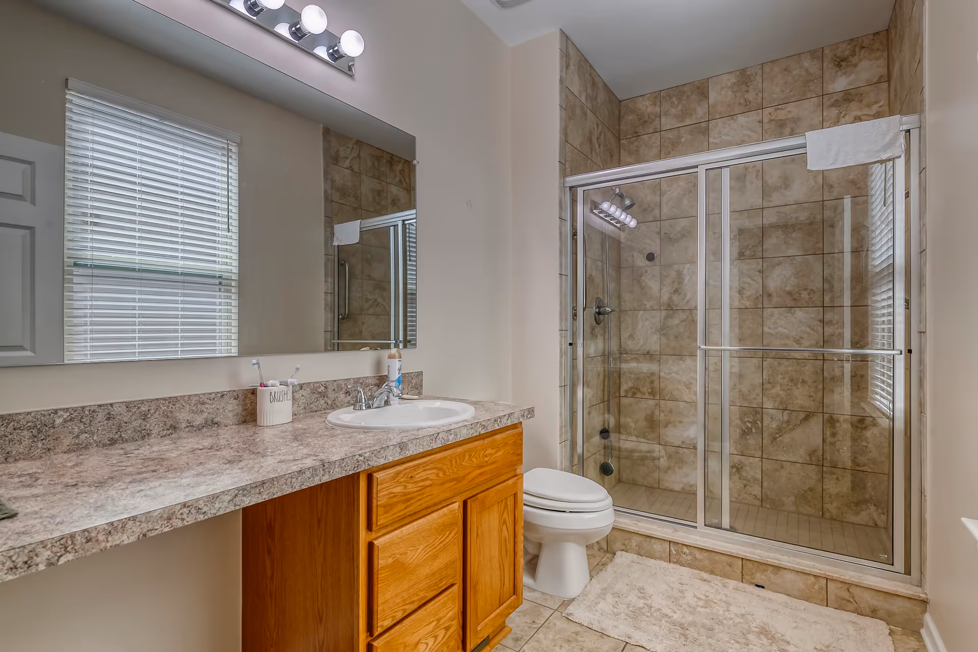 A bathroom with a large mirror above a countertop with a sink and wooden cabinets below. There is a toothbrush holder and a soap dispenser on the countertop. Next to the sink is a toilet, and beside it is a glass-enclosed shower with beige tiled walls and a white towel hanging on the shower door. A window with blinds is visible on the left wall, and a white bath mat is on the tiled floor.