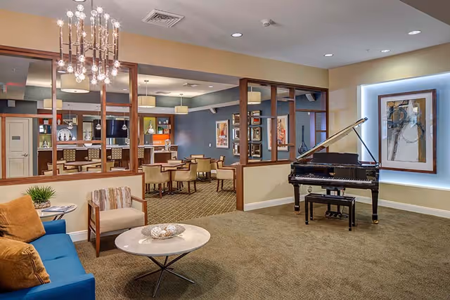 A spacious senior living common area featuring a blue couch with orange pillows, a beige armchair, and a round marble coffee table. A black grand piano is positioned near a wall with a framed abstract painting illuminated by soft lighting. In the background, there is a dining area with tables and chairs, separated by wooden-framed glass partitions. The room is well-lit with ceiling lights and a modern chandelier.