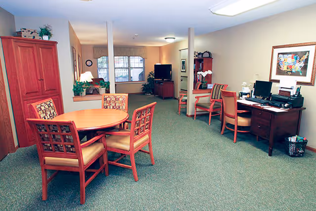 A spacious interior room with green carpet and beige walls, featuring a round wooden table with four cushioned chairs in the foreground. To the right, there is a wooden desk with a computer, printer, and office supplies, accompanied by two chairs. In the background, a living area with a TV on a stand, a red cabinet, and a window with blinds is visible. The room is softly lit with ceiling lights and a table lamp.