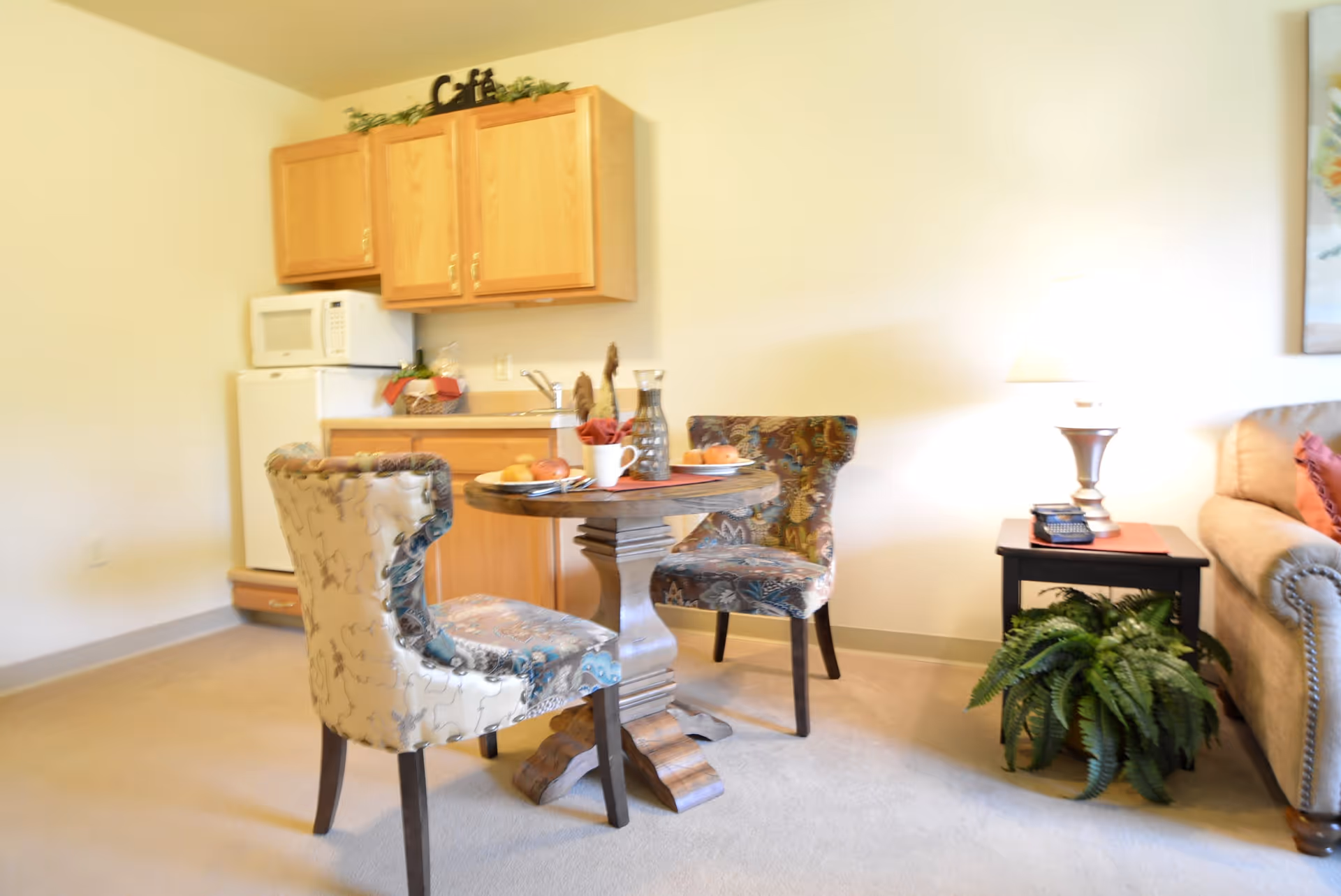 A cozy assisted living area featuring a small round wooden table with two upholstered chairs, a kitchenette with wooden cabinets, a microwave, and a mini refrigerator. To the right, there is a beige sofa, a side table with a lamp, a phone, and a green potted plant underneath.