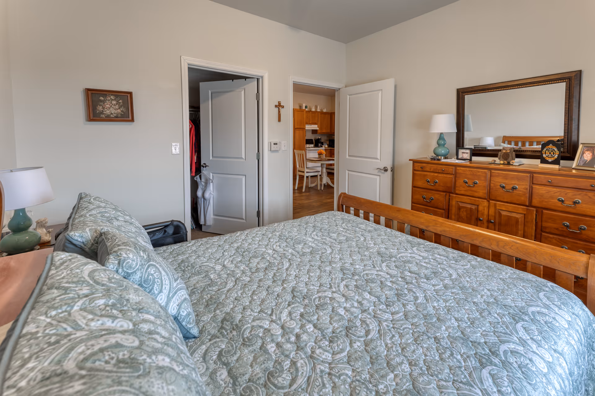 A bedroom with a wooden bed frame and a green patterned quilt. There are matching pillows on the bed. To the right is a wooden dresser with a large mirror, a lamp, and framed photos. Two doors are visible, one leading to a closet and the other to a kitchen area with a dining table and chairs. The walls are light-colored and there is a small framed picture and a cross hanging on the wall.
