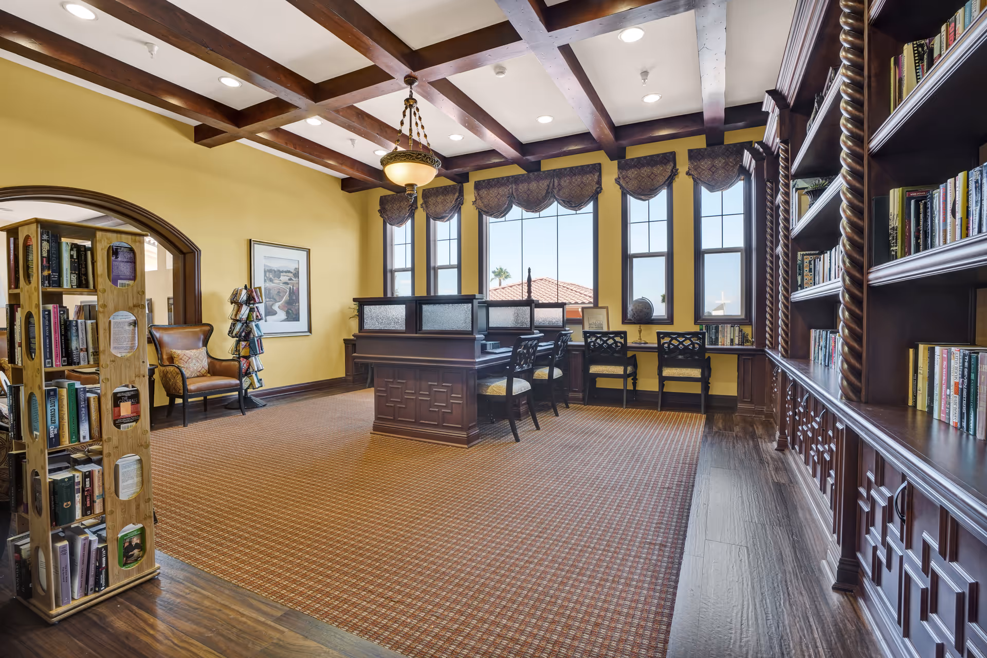 A cozy library room with yellow walls and large windows letting in natural light. The room features wooden bookshelves filled with books, a wooden book display rack, a comfortable armchair, and a desk with chairs facing the windows. The ceiling has wooden beams and a hanging light fixture.