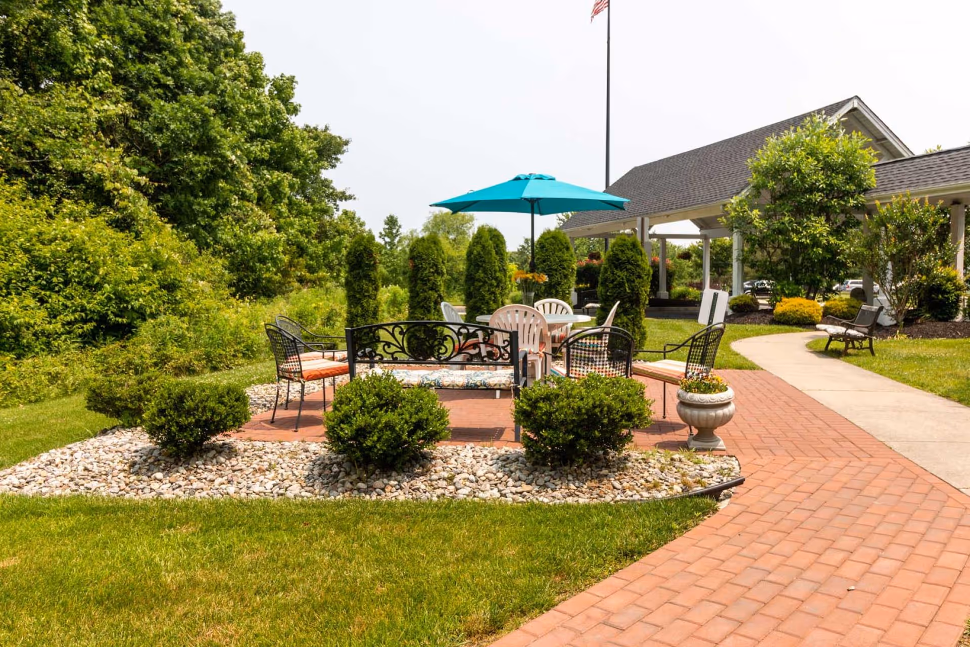 Outdoor seating area at Voorhees Senior Living featuring a brick patio with metal and plastic chairs, a table with a blue umbrella, surrounded by green bushes and trees, with a covered walkway and building in the background.