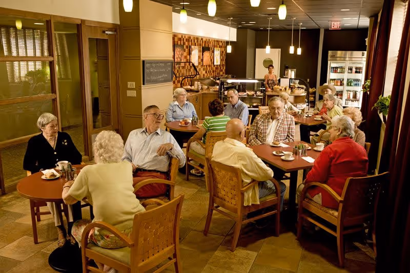 A group of elderly people sitting at round tables in a cozy dining area, engaging in conversation and enjoying beverages. The room has warm lighting with pendant lights hanging from the ceiling, a counter with food and drinks in the background, and large windows with curtains on the right side.