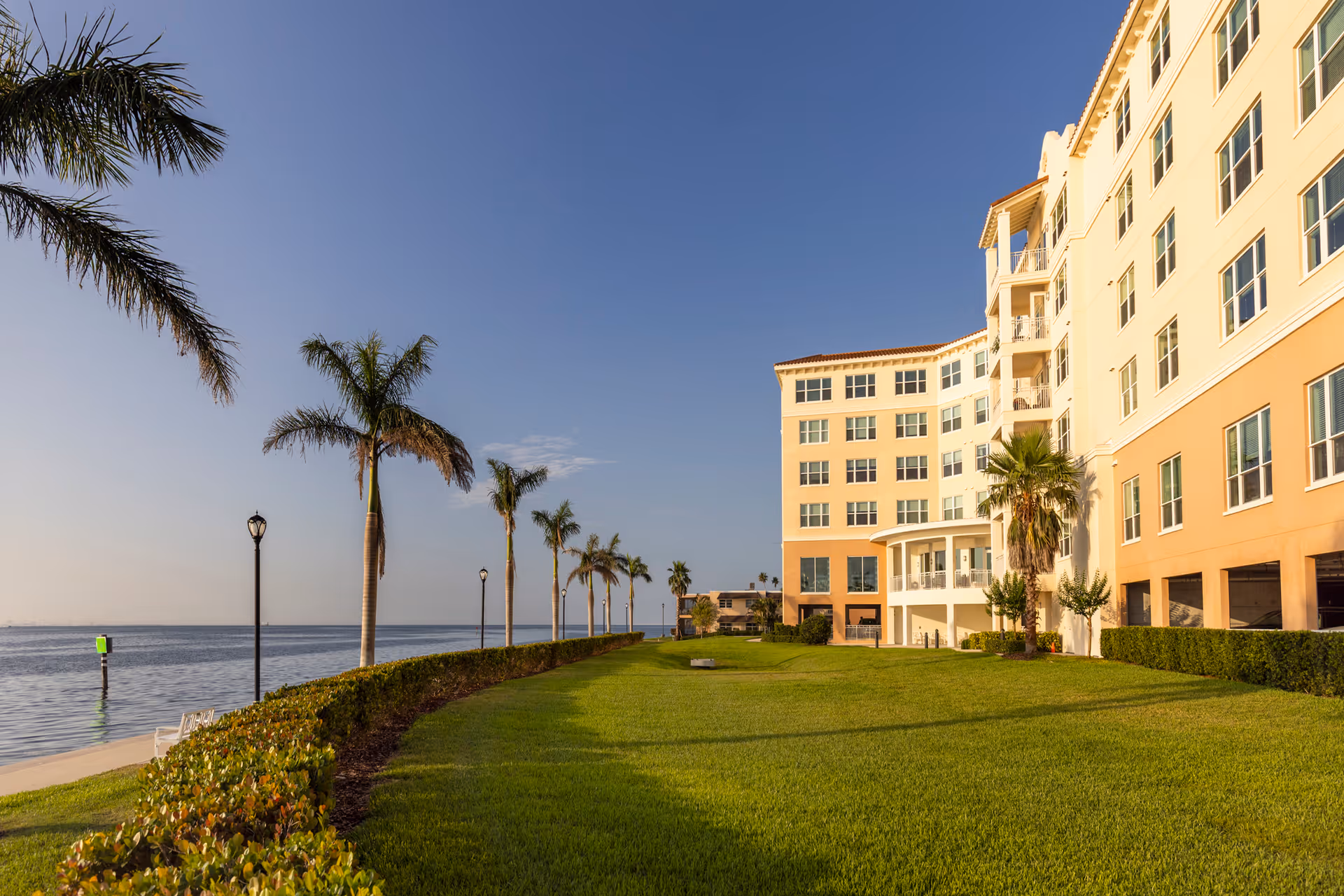 A waterfront view of a multi-story senior living facility building with a well-maintained lawn, palm trees, and a clear blue sky.