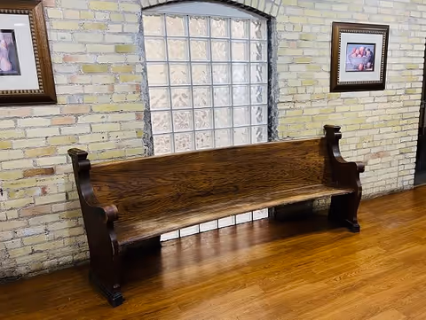 A long wooden bench with carved armrests placed against a light yellow brick wall with a large glass block window in the center. Two framed pictures hang on the wall, one on each side of the window. The floor is made of polished wood.