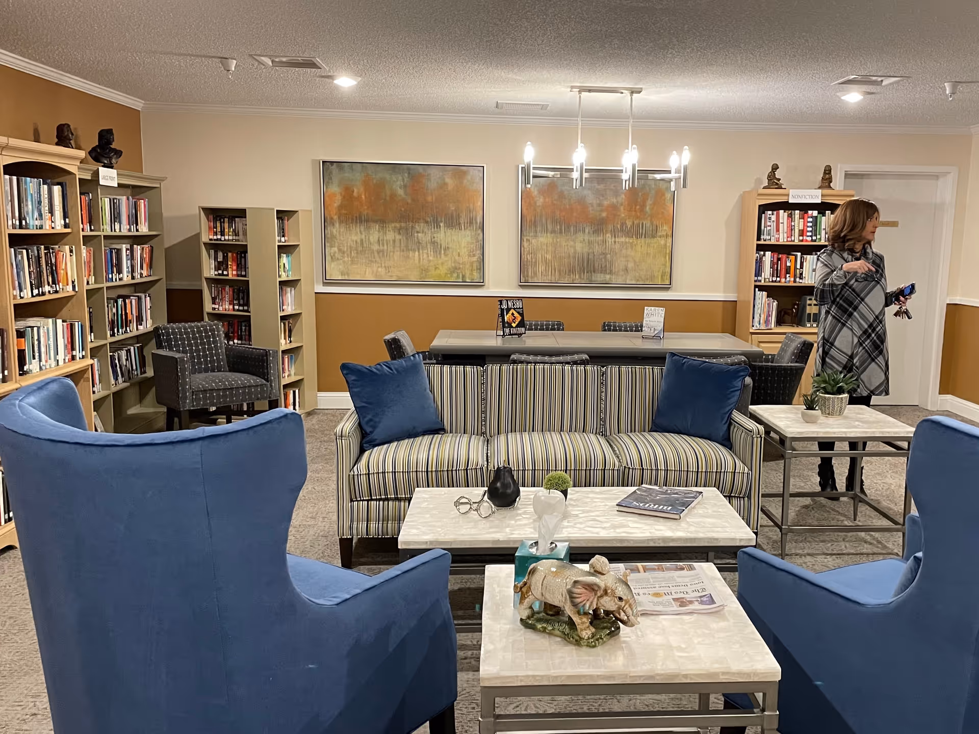 A well-lit community lounge with blue armchairs, a striped sofa, coffee tables and bookshelves.