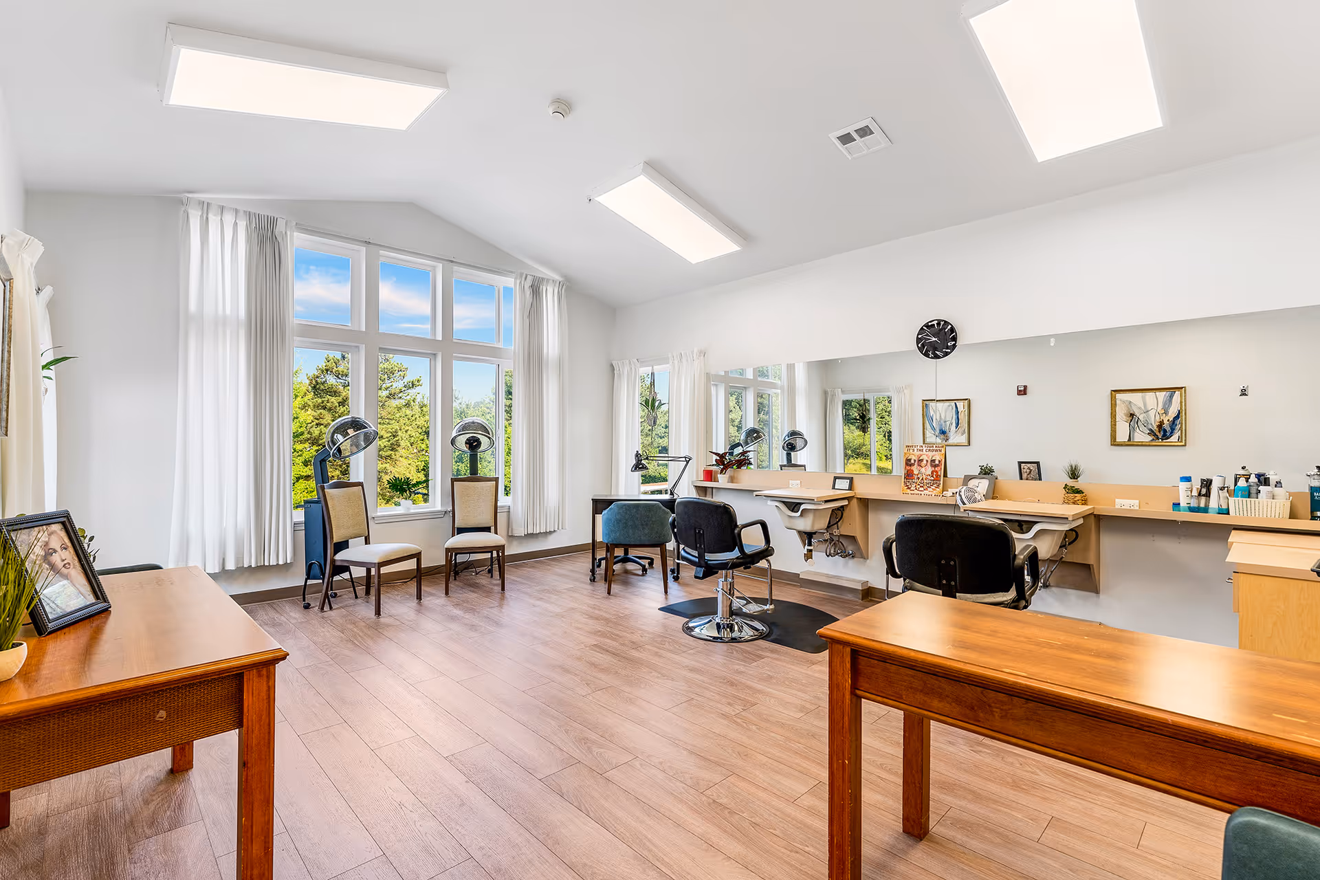A bright and spacious hair salon area with large windows letting in natural light. The room features wooden flooring, two hair styling chairs in front of a long mirror with hair care products on the counter, and two waiting chairs near the windows. The walls are white with minimal decorations, including framed artwork and a clock.