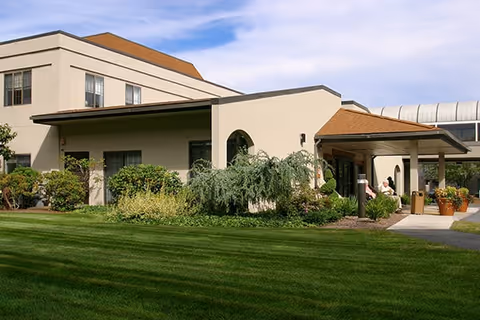 Exterior view of a beige multi-story building with a covered entrance, surrounded by green grass, shrubs, and plants under a partly cloudy sky.