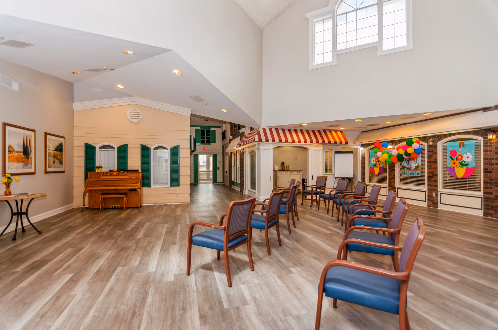 A spacious senior living community common area with wooden flooring, multiple chairs arranged in rows, a piano against the wall, colorful balloons, and decorative window displays with flower designs and a welcome sign. The room has high ceilings with windows near the top and soft lighting.