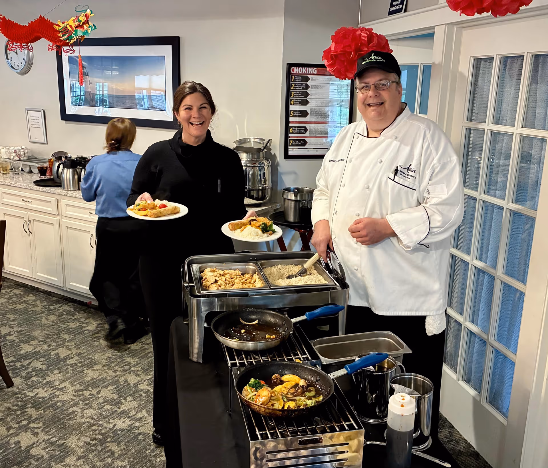 A smiling chef in a white uniform and black cap stands behind a buffet table with pans of cooked food. A woman dressed in black holds two plates of food and smiles at the camera. Another person in a blue shirt is seen in the background near a counter with coffee pots and other items. The setting appears to be a dining or serving area inside a facility.