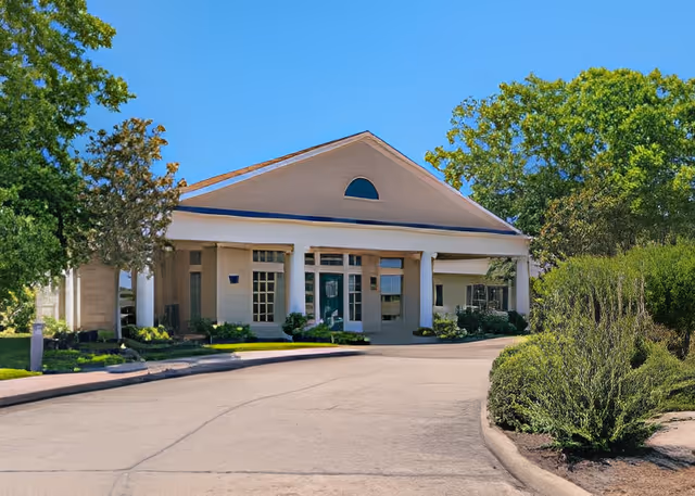 Front entrance of a single-story assisted living building with columns, a circular driveway, and landscaped shrubs.