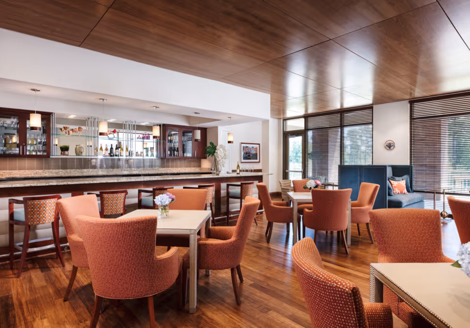 A bright and modern dining area with several square tables surrounded by orange upholstered chairs. In the background, there is a bar with high stools, glass shelves displaying bottles and glassware, and pendant lights hanging from the ceiling. Large windows with wooden blinds allow natural light to fill the room, and a blue cushioned bench with decorative pillows is visible near the windows.