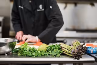 A person wearing a black chef's jacket is chopping vegetables on a kitchen counter. Fresh vegetables including cilantro, a lime, and asparagus are laid out on the counter along with a metal mixing bowl.