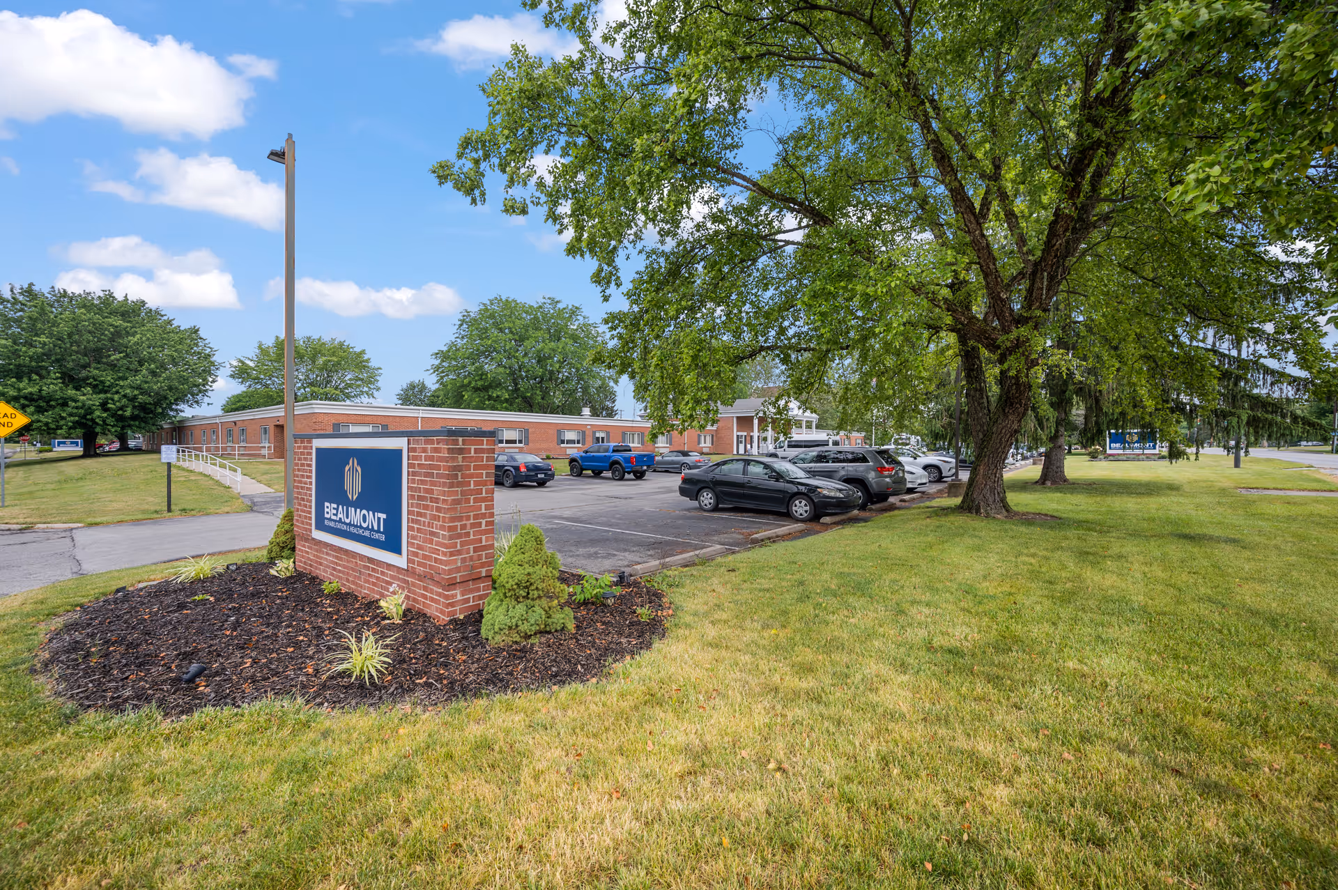 Exterior view of Beaumont Rehabilitation and Healthcare Center showing a brick sign with the facility name, a parking lot with several cars, green grass, large trees, and a partly cloudy blue sky.