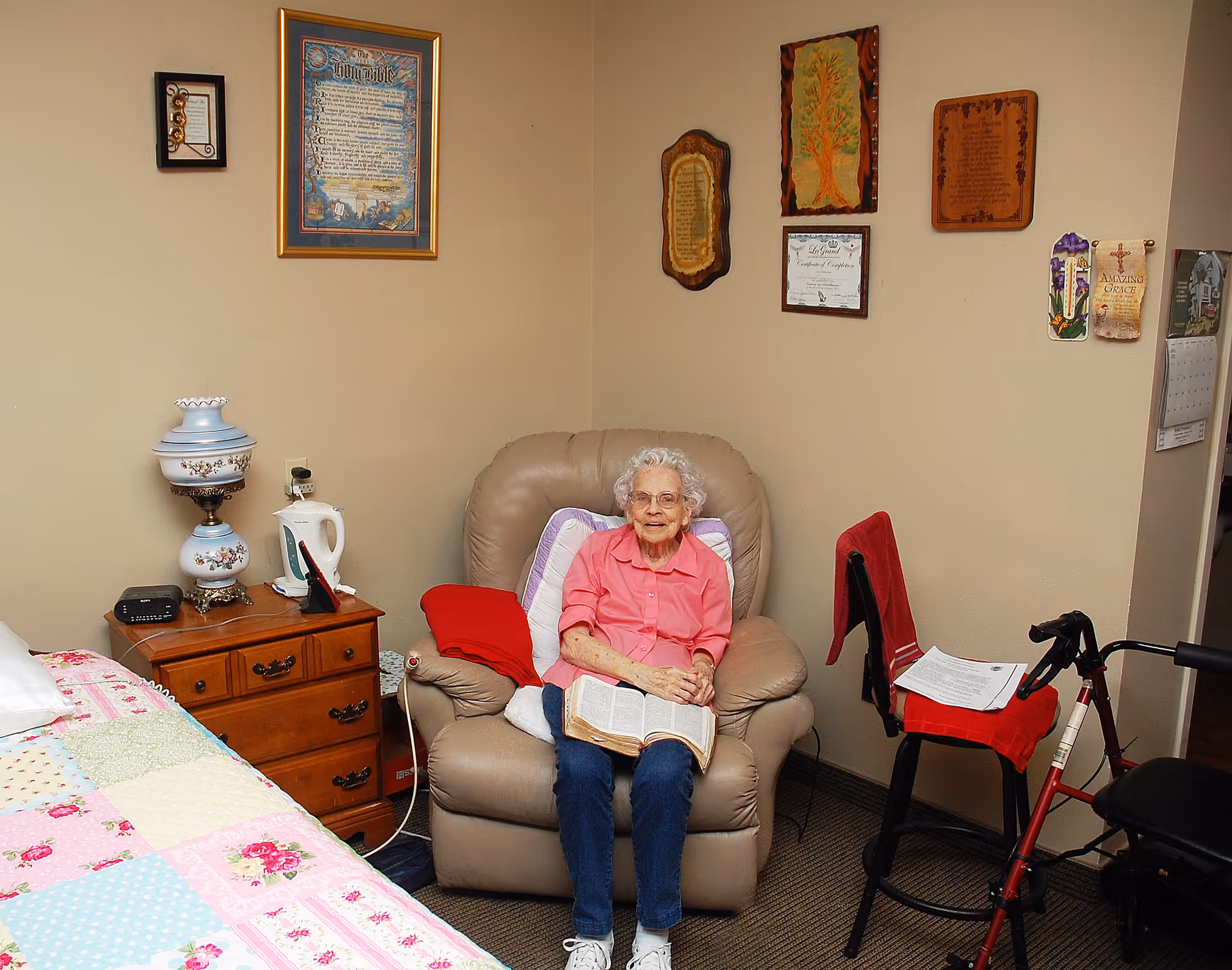 An elderly woman with white hair and glasses sits in a beige recliner chair in a cozy room. She is wearing a pink shirt and blue jeans and holds an open book on her lap. The room has a wooden nightstand with a decorative lamp and an electric kettle. The wall behind her is decorated with framed certificates, plaques, and artwork. A bed with a colorful patchwork quilt is partially visible on the left, and a red walker with a seat and papers on it is on the right.