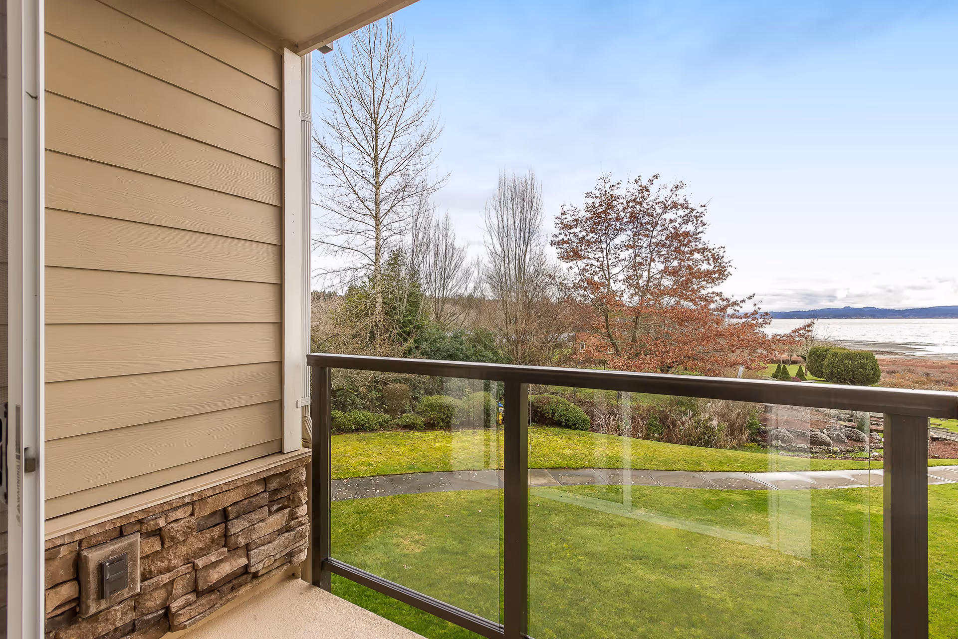 View from a balcony with a glass railing overlooking a green lawn, trees with some autumn leaves, and a body of water in the distance under a partly cloudy sky.