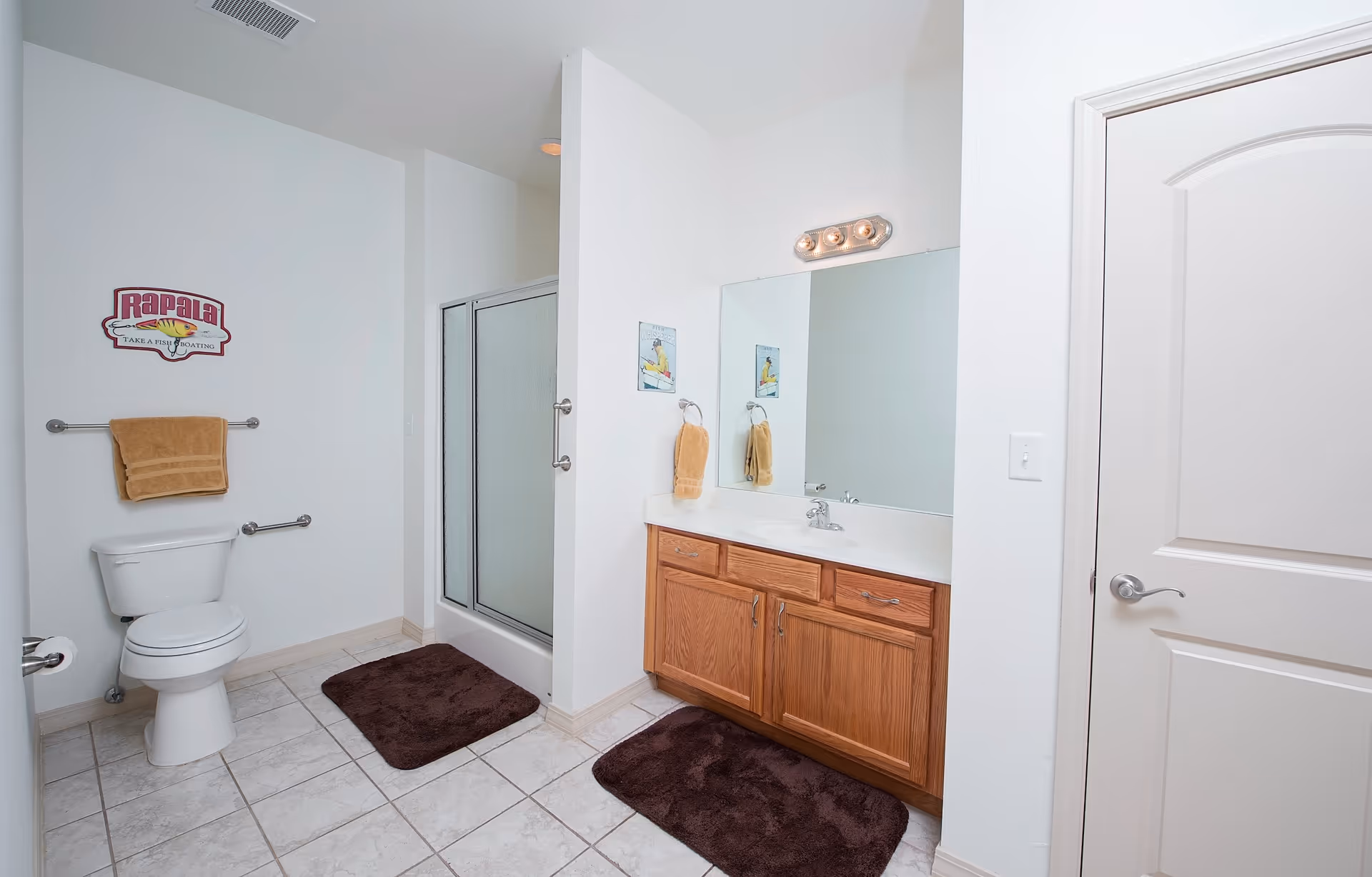 A bright bathroom featuring a toilet, glass-enclosed shower, wooden vanity with sink and mirror, and brown bath mats.