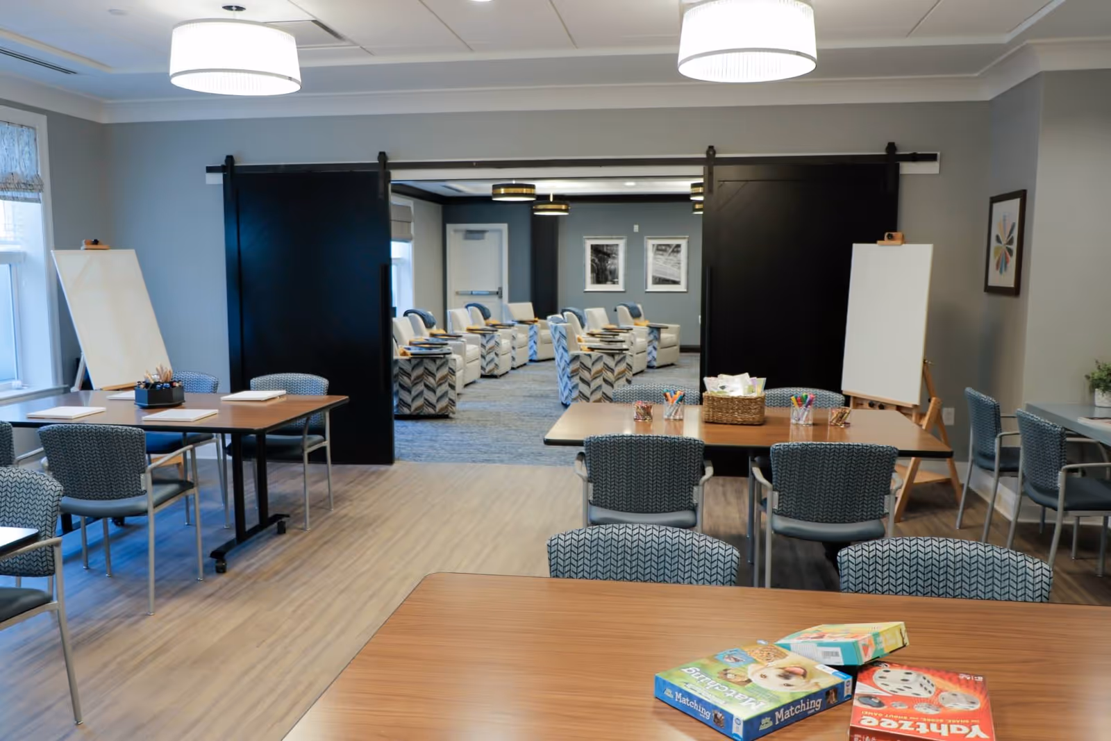 Bright communal activity room with tables, chairs, art easels and craft supplies looking through sliding barn doors into a seating area.