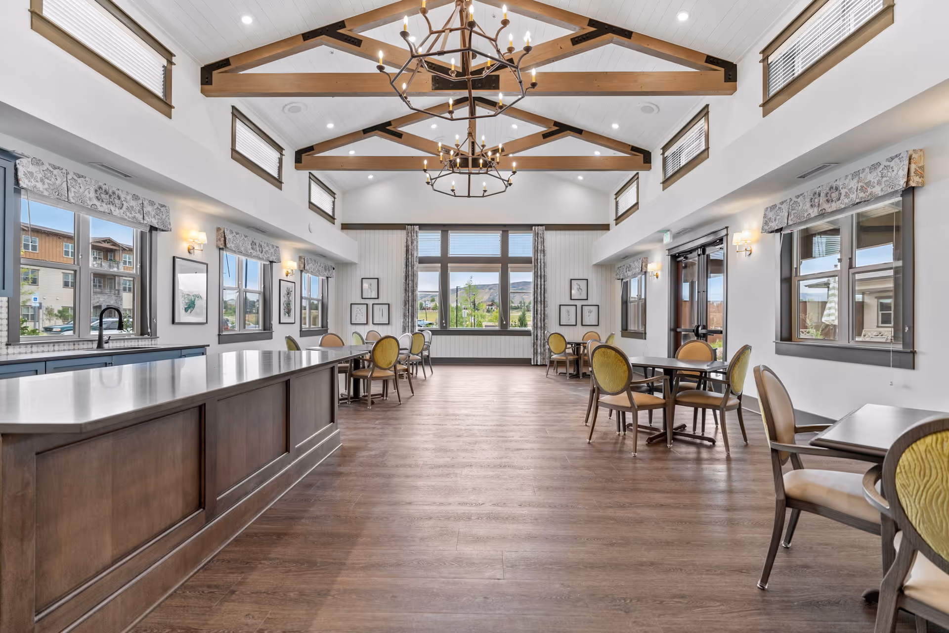 Spacious dining room with wooden floors, multiple round tables with chairs, large windows allowing natural light, and a high ceiling with exposed wooden beams and chandeliers. There is a long kitchen island on the left side with a sink and cabinetry underneath.