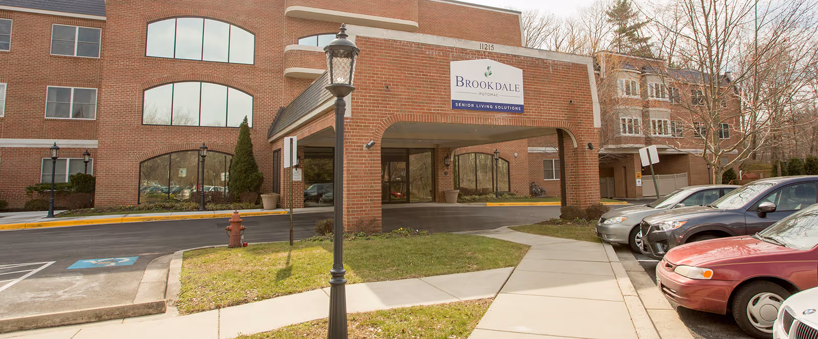 Exterior view of the Brookdale Potomac senior living facility showing a brick building with large windows, a covered entrance with a sign reading 'Brookdale Potomac Senior Living Solutions,' a sidewalk, parked cars, and a street lamp.