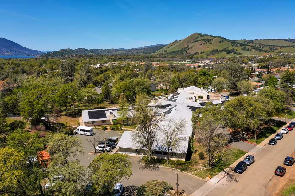 Aerial view of a single-story nursing center complex surrounded by trees with rolling hills and a lake visible in the distance.